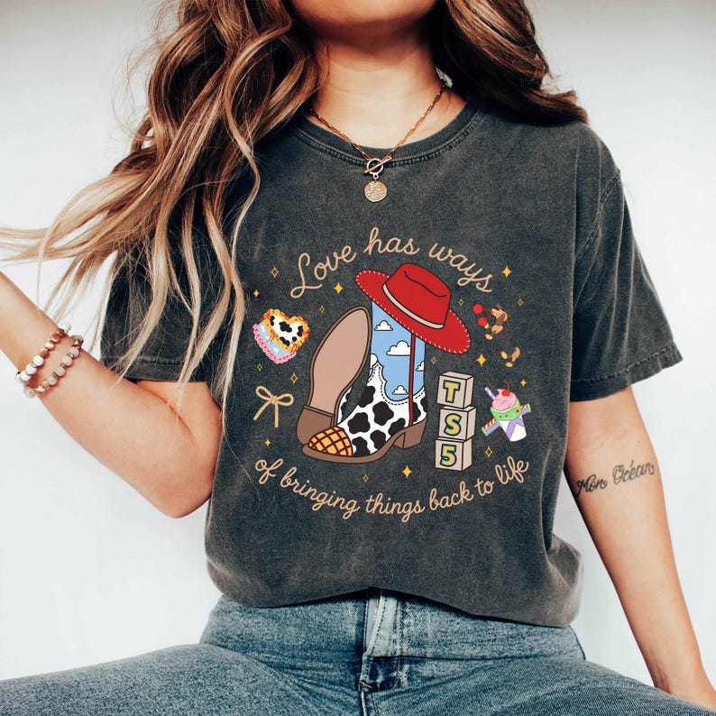 May include: A charcoal gray short-sleeve t-shirt featuring a graphic print of a red cowboy hat, a cow-print boot, alphabet blocks spelling TSS, and a milkshake. The text reads Love has ways of bringing things back to life in cursive script.