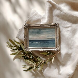 May include: A framed seascape painting with a silver ornate frame. The artwork depicts a blue ocean under a cloudy sky. A sprig of green leaves rests near the frame, set against a neutral fabric background.