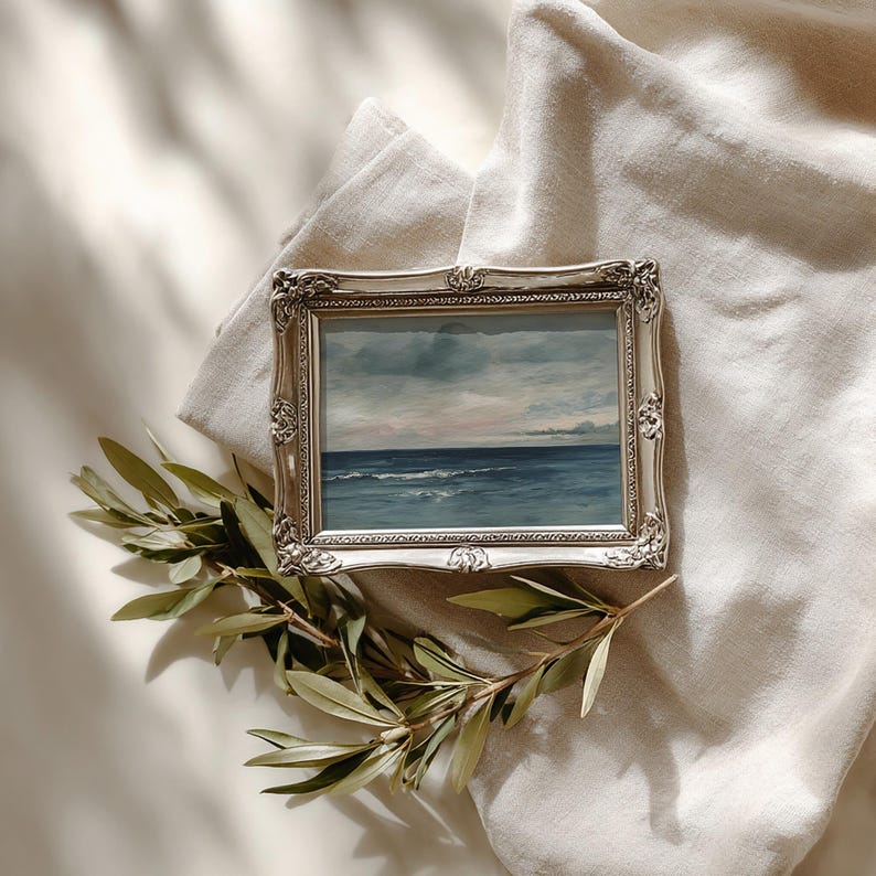 May include: A framed seascape painting with a silver ornate frame. The artwork depicts a blue ocean under a cloudy sky. A sprig of green leaves rests near the frame, set against a neutral fabric background.
