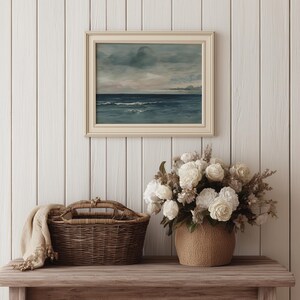May include: A framed seascape painting with a cream-colored frame hangs above a wooden table. A woven basket with a handle and a beige throw blanket sit on the table. A bouquet of white flowers in a woven pot completes the scene.