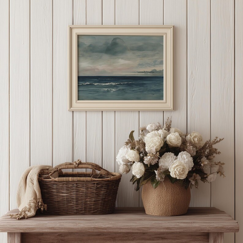 May include: A framed seascape painting with a cream-colored frame hangs above a wooden table. A woven basket with a handle and a beige throw blanket sit on the table. A bouquet of white flowers in a woven pot completes the scene.