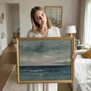 May include: A framed landscape painting depicting a seascape with a cloudy sky. The artwork features shades of blue, gray, and hints of pink. The ornate gold frame adds a classic touch to the piece, suitable for home decor.