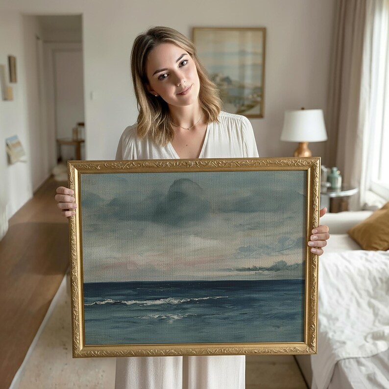 May include: A framed landscape painting depicting a seascape with a cloudy sky. The artwork features shades of blue, gray, and hints of pink. The ornate gold frame adds a classic touch to the piece, suitable for home decor.