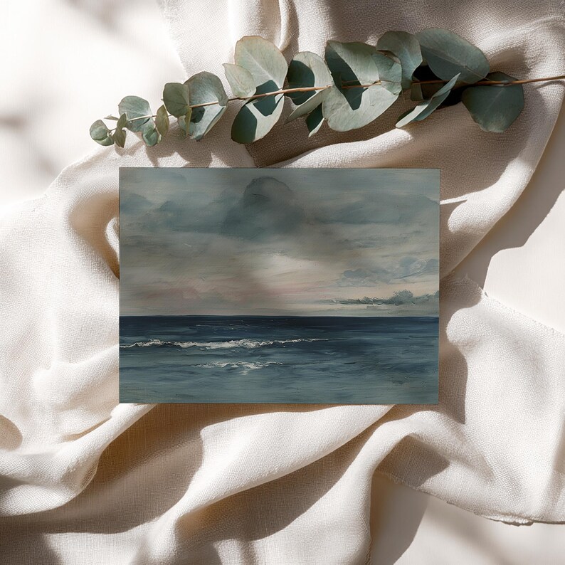 May include: An art print depicting a seascape with a moody sky and ocean. The painting features shades of blue, gray, and white, with visible brushstrokes. The artwork is displayed on a white surface with a sprig of eucalyptus.