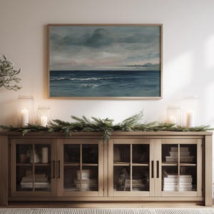 May include: A wooden cabinet with glass doors displays books and decorative items. Above it, a framed seascape painting hangs, depicting a cloudy sky and ocean waves. Lit candles in glass lanterns and greenery add to the room's ambiance.