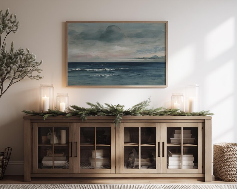 May include: A wooden cabinet with glass doors displays books and decorative items. Above it, a framed seascape painting hangs, depicting a cloudy sky and ocean waves. Lit candles in glass lanterns and greenery add to the room's ambiance.