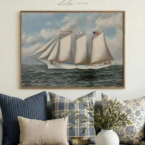 May include: A framed nautical painting of a white schooner sailing on a wavy sea under a cloudy sky. The painting has three white sails and flags. The frame is a light brown color. The painting is above a gray couch with blue pillows.