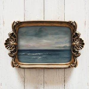 May include: A rectangular, gold-colored decorative tray with ornate scrollwork and handles. The tray holds a painting of a seascape with a dark blue ocean, white waves, and a cloudy sky with hints of pink.