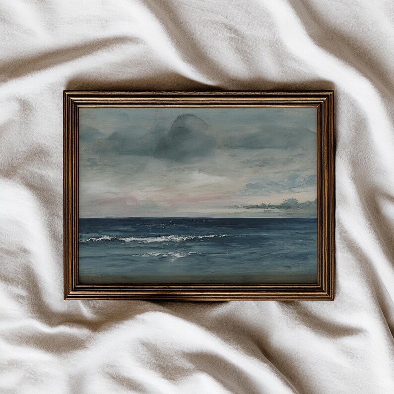 May include: A framed seascape painting depicts a moody ocean scene. The artwork features a dark blue sea with white-capped waves under a cloudy sky with hints of pink and gray. The frame is a warm, golden brown.