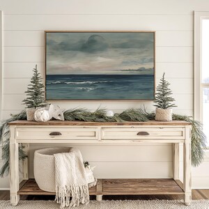 May include: A cream-colored console table with two drawers and a lower shelf, decorated for the holidays. A framed seascape painting hangs above the table. Two small Christmas trees in woven pots flank the painting, with a garland and decorative accents.