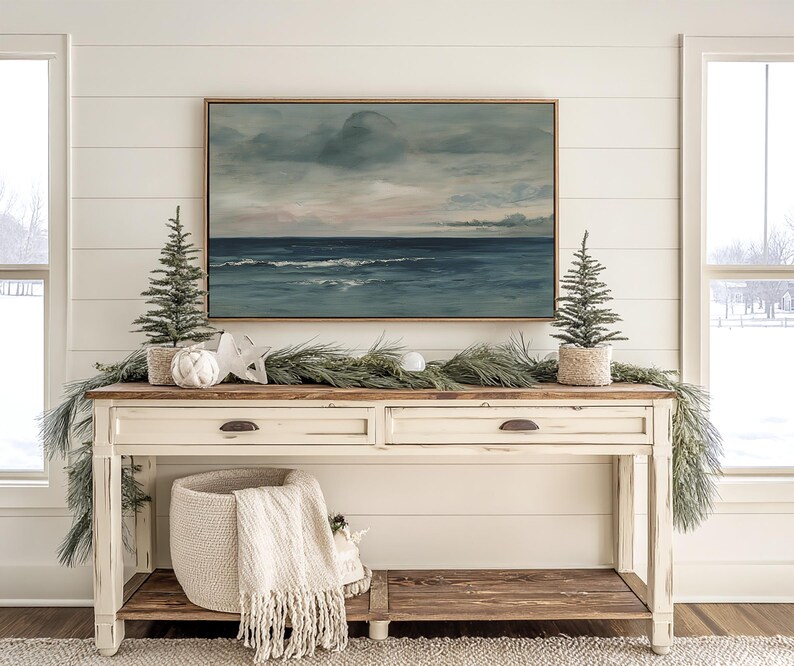 May include: A cream-colored console table with two drawers and a lower shelf, decorated for the holidays. A framed seascape painting hangs above the table. Two small Christmas trees in woven pots flank the painting, with a garland and decorative accents.