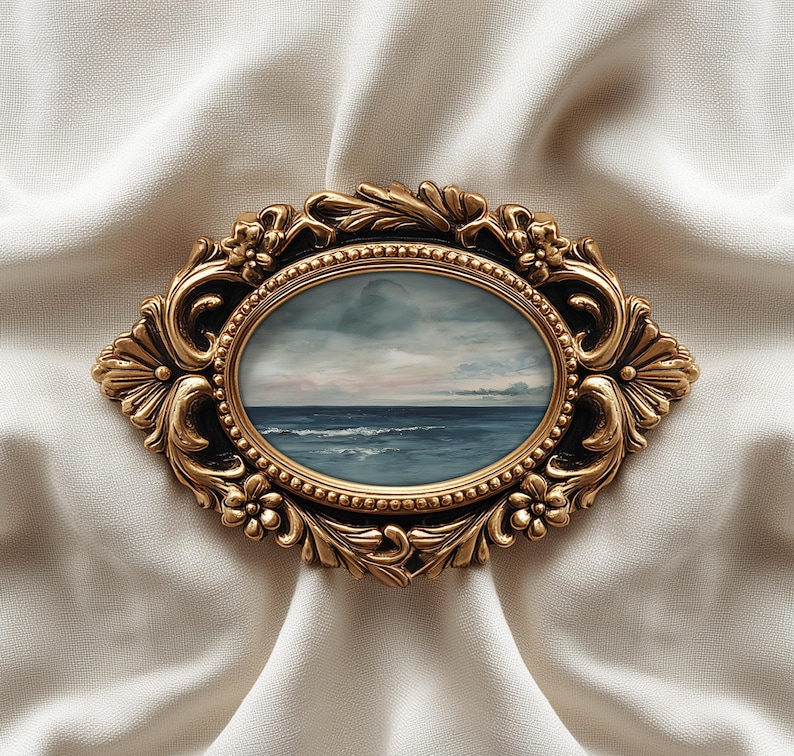 May include: An oval-shaped, antique gold-colored frame with an ocean landscape painting. The frame has ornate floral and scrollwork details. The painting depicts a seascape with a cloudy sky and blue water. The frame is set against a white, textured fabric.