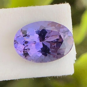 May include: An oval-cut gemstone with a vibrant purple hue and multifaceted surface, displaying internal light reflections and deep violet tones. The stone is positioned against a plain white rectangular background, highlighting its clarity and colour.