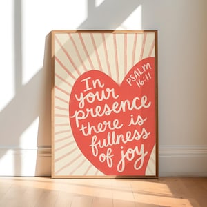 May include: A framed rectangular wall art print featuring a large red heart shape against a cream background with radiating light-colored lines. The text reads: In your presence there is fullness of joy Psalm 16:11 in white cursive lettering.