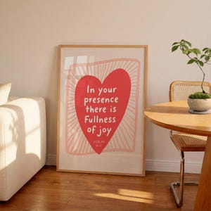 May include: A rectangular framed wall art print featuring a large red heart centred on a light background with radiating lines. The text inside the heart reads: In your presence there is Fullness of joy PSALM 16:11. The print is displayed in a light wood frame.