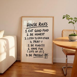 May include: A framed print with the words "HOUSE RULES" in a handwritten style. The rules are listed below the title, including "EAT GOOD FOOD," "BE HONEST," and "BE PRESENT." The frame is a light wood color, and the background is white.