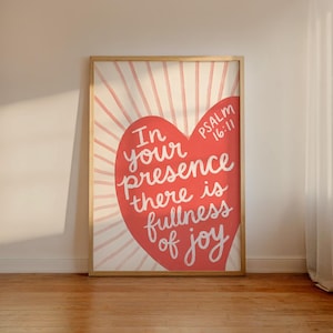 Puede incluir: Un cuadro rectangular con marco que presenta un gran corazón rojo centrado sobre un fondo crema con líneas de rayos de sol en rosa claro. El texto dice In your presence there is fullness of joy Psalm 16:11 en letras cursivas blancas.