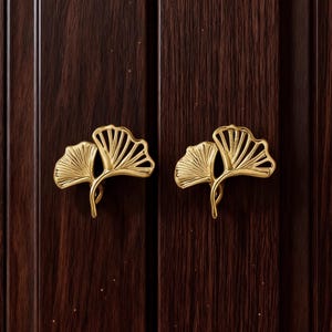 May include: Two gold-coloured cabinet pulls shaped like ginkgo leaves are mounted on a dark brown wooden cabinet door. The pulls have detailed veining and a curved stem. The cabinet door has vertical wood grain.
