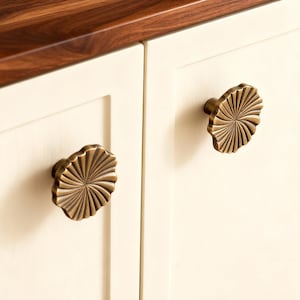 Może przedstawiać: Zbliżenie na dwoje kremowych drzwi szafki z dekoracyjnymi, brązowymi gałkami. Gałki mają wzór promieni słonecznych. Ciemnobrązowy drewniany blat jest widoczny nad szafkami.