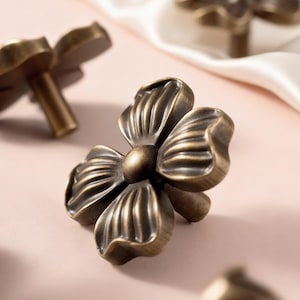 May include: Close-up of several antique brass flower-shaped cabinet knobs. Each knob features four detailed petals and a central rounded accent. The knobs are arranged on a soft pink surface, with a white satin fabric in the background.