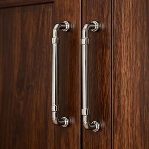 May include: Close-up of two polished chrome cabinet handles against a dark brown wood grain cabinet door. The handles are cylindrical with curved ends and decorative circular accents. The wood grain has a rich, warm tone.
