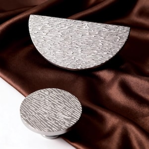 May include: Two silver-toned cabinet hardware pieces. One is a semi-circular handle, and the other is a round knob. Both have a textured surface. The hardware is displayed on a brown satin fabric.