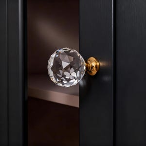 May include: A faceted crystal ball cabinet knob with a gold-colored base is mounted on a black cabinet door. The knob reflects light, creating a sparkling effect. The cabinet door is dark, and the background is blurred.