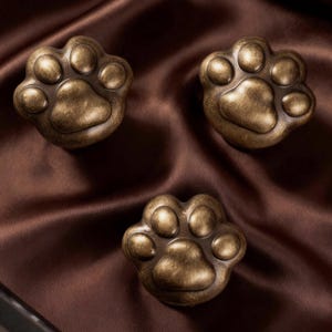 May include: Three bronze-colored paw print cabinet knobs. Each knob features a detailed paw pad and toe design. The knobs are arranged on a brown satin fabric, creating a warm and inviting aesthetic. These knobs are ideal for adding a unique touch to furniture.