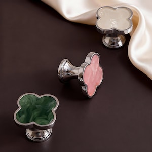 May include: Three decorative cabinet knobs with a cloverleaf design. The knobs have a polished silver base and a clover-shaped top in white, pink, and green. The knobs are set against a dark brown background with a white satin fabric.