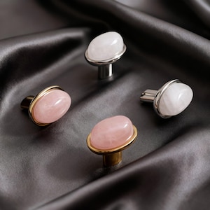 May include: Four cabinet knobs with oval rose quartz stones. Two knobs have gold-toned bases, and two have silver-toned bases. The rose quartz stones are a soft pink colour. The knobs are set against a dark grey satin background.