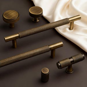 May include: An assortment of antique brass cabinet hardware, including knobs and pulls. The knobs are round with a textured surface, and the pulls are bar-shaped with a diamond pattern. The hardware is displayed on a dark brown surface with a white satin fabric.