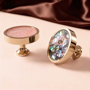 Può includere: Due pomelli decorativi per mobili. Uno presenta una parte superiore circolare rosa con una base color oro. L'altro ha una forma ovale con intarsi di conchiglia di abalone iridescente e una base color oro. Entrambi i pomelli sono su una superficie rosa chiaro.