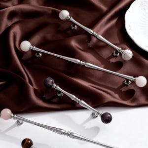 May include: A collection of polished silver cabinet pulls with decorative gemstone sphere ends. The pulls are displayed on a brown satin fabric background. The gemstones include white, purple, and pink, adding a touch of elegance to the hardware.