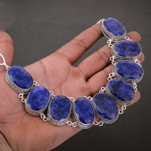 Può includere: Una collana d'argento con grandi pietre preziose ovali blu scuro. Le pietre sono incastonate in castoni d'argento e collegate da maglie d'argento. La collana è mostrata su uno sfondo scuro.