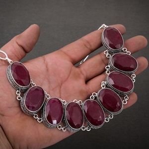 May include: A silver necklace featuring large, oval-shaped, deep red gemstones. The gemstones are set in ornate silver bezels and linked together with delicate silver chain. The necklace is displayed on a hand against a dark background.