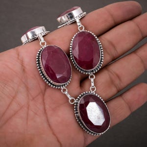 Könnte beinhalten: Eine silberne Halskette mit drei großen, oval geformten, tiefroten Rubin-Edelsteinen. Die Rubine sind in verzierten Silberfassungen mit einem Perlenrand gefasst. Die Halskette hat ein klassisches, elegantes Design.