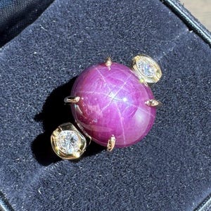 May include: A gold ring featuring a large, round, deep pink gemstone, possibly a star ruby, is presented in a black velvet jewellery box. Two smaller, diamond-set gold accents flank the main stone, adding sparkle and contrast.