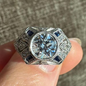 K&ouml;nnte beinhalten: Ein silberner Ring mit einem gro&szlig;en, runden, klaren Edelstein in der Mitte, umgeben von kleineren klaren Steinen und blauen rechteckigen Steinen. Der Ring hat ein aufwendiges Design im Art-Deco-Stil.