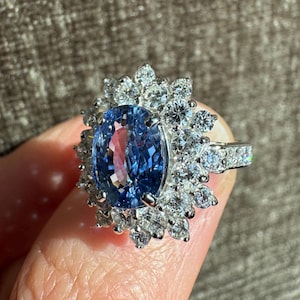 May include: A dazzling ring featuring a large, oval-cut blue gemstone surrounded by a halo of smaller, clear gemstones. The ring's band is also adorned with clear gemstones, creating a sparkling and elegant design.