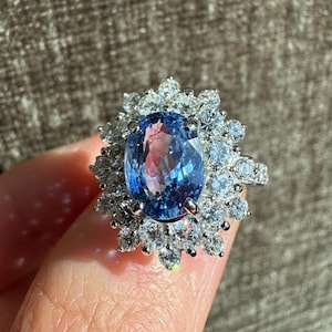 May include: A silver ring featuring a large, oval-cut blue gemstone surrounded by smaller, clear gemstones. The ring's design is a cluster of sparkling stones around the central blue gem, creating a halo effect. The ring is held up against a neutral background.