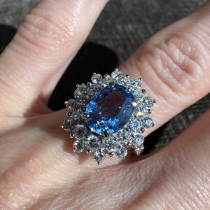May include: A silver ring featuring a large, oval-cut blue gemstone surrounded by smaller, clear gemstones. The ring's design is a cluster of stones, creating a sparkling, eye-catching effect. The ring is shown on a person's finger.