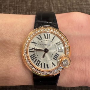 Puede incluir: Un reloj de pulsera dorado con esfera plateada y n&uacute;meros romanos negros. El reloj presenta un bisel con incrustaciones de diamantes y una correa de cuero negro. La palabra "CARTIER" est&aacute; impresa en la esfera. Hecho en Suiza.