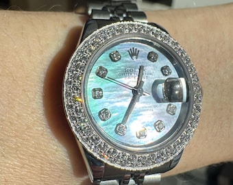 Può includere: Un orologio da polso argentato con quadrante in madreperla e lunetta tempestata di diamanti. L'orologio presenta un datario e un cinturino argentato. L'orologio è al polso.