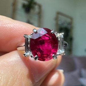May include: A ring featuring a large, oval-cut, deep red ruby gemstone set in a silver-toned metal band. The central ruby is flanked on each side by a single, rectangular, baguette-cut clear diamond, secured by metal prongs in a three-stone design.