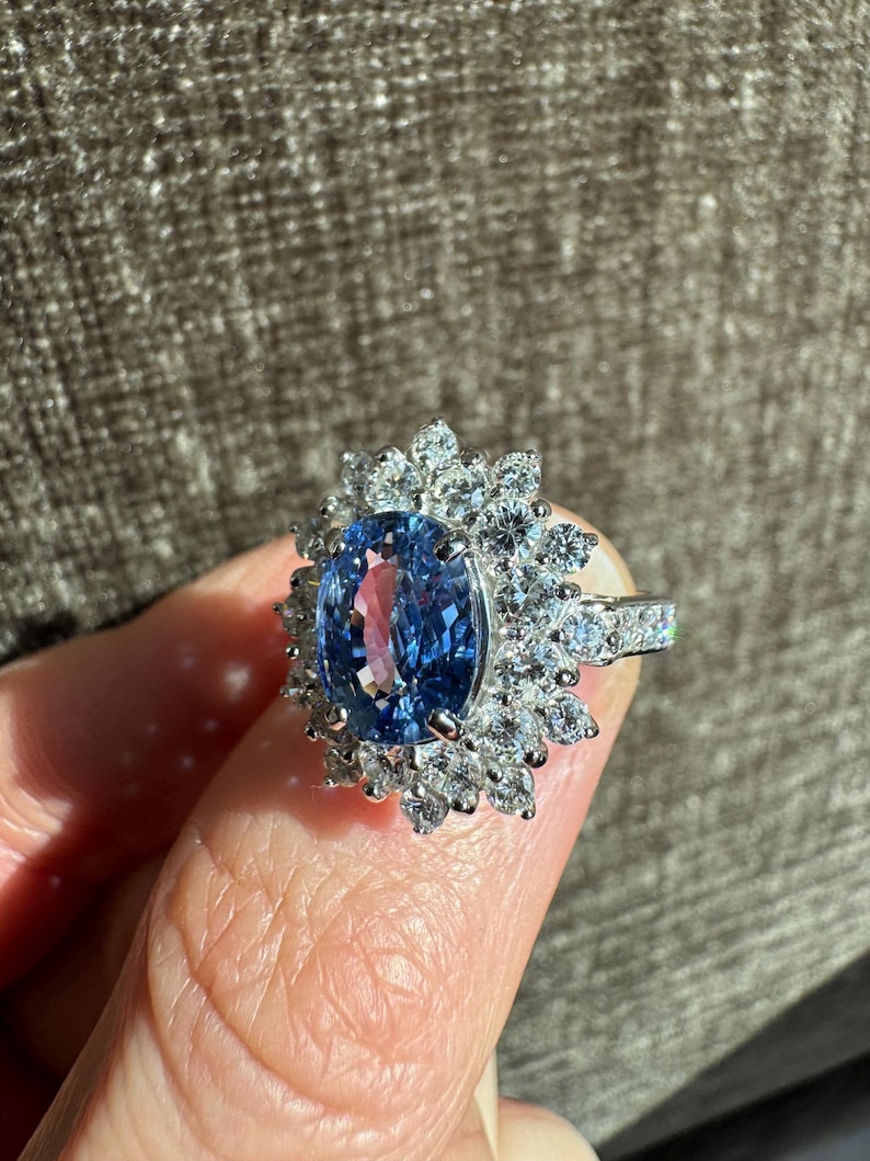 May include: A dazzling ring featuring a large, oval-cut blue gemstone surrounded by a halo of smaller, clear gemstones. The ring's band is also adorned with clear gemstones, creating a sparkling and elegant design.