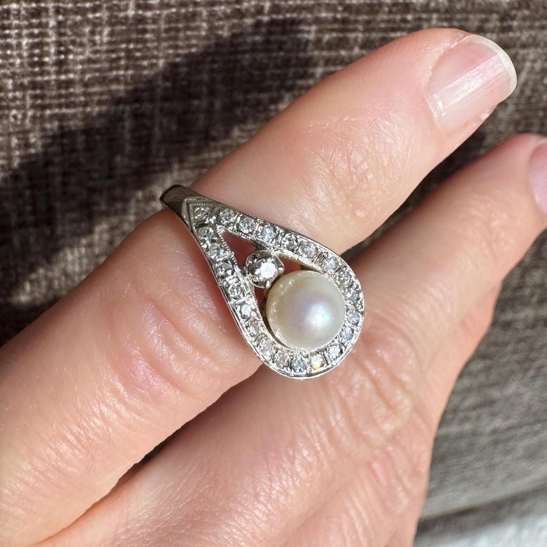 May include: A silver ring with a pear-shaped design, featuring a central pearl and surrounding clear gemstones. The ring is set against a neutral background, highlighting its elegant details.