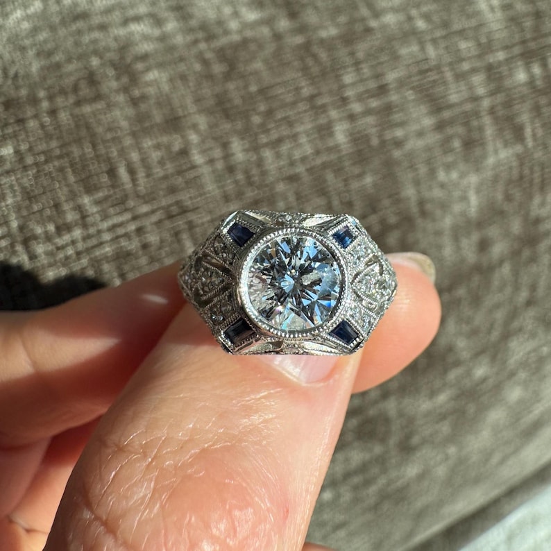 K&ouml;nnte beinhalten: Ein silberner Ring mit einem gro&szlig;en, runden, klaren Edelstein in der Mitte, umgeben von kleineren klaren Steinen und blauen rechteckigen Steinen. Der Ring hat ein aufwendiges Design im Art-Deco-Stil.