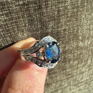 May include: A silver ring with a large, oval, deep blue gemstone as the centerpiece. The ring is encrusted with smaller, clear gemstones around the main stone and along the band. The setting has intricate details, and the ring is held up by a finger.