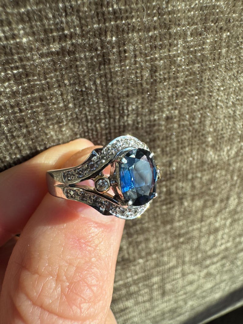May include: A silver ring with a large, oval, deep blue gemstone as the centerpiece. The ring is encrusted with smaller, clear gemstones around the main stone and along the band. The setting has intricate details, and the ring is held up by a finger.