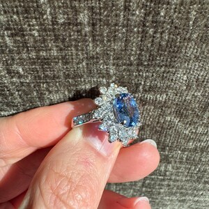 May include: A silver ring featuring a large, oval-cut blue gemstone surrounded by smaller, clear gemstones. The ring's band is also adorned with smaller, clear gemstones. The ring is held up against a textured, brown background.